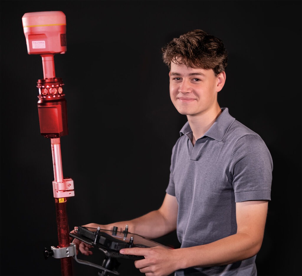 Ein junger Mann in einem grauen Polohemd hält eine Fernbedienung in der Hand und steht neben einem Vermessungsinstrument, vor einem dunklen Hintergrund.