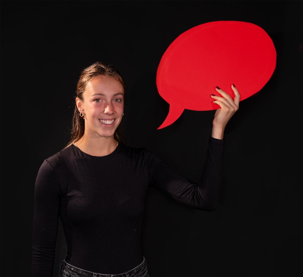 Eine Person in einem schwarzen Oberteil hält einen großen roten Sprechblasen-Ausschnitt in der Hand vor einem schwarzen Hintergrund