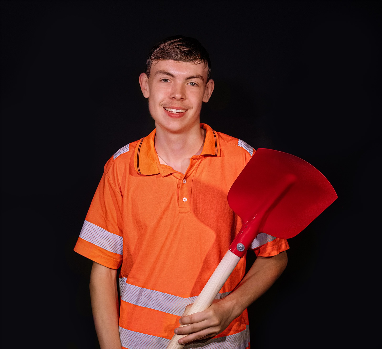 Ein junger Mann in einem orangefarbenen Warnschutzshirt hält eine rote Schaufel vor einem schwarzen Hintergrund und lächelt in die Kamera.