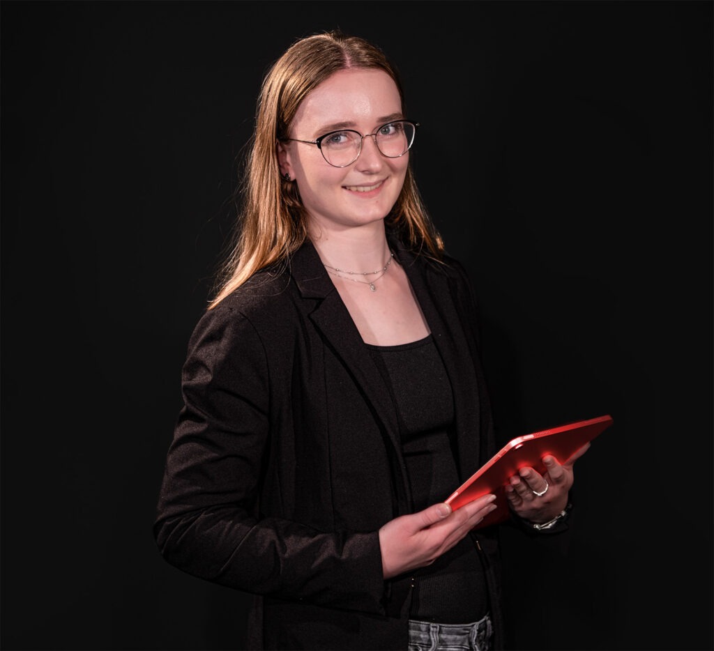 Eine lächelnde junge Frau mit Brille und langen, blonden Haaren hält ein rotes Tablet und trägt eine schwarze Jacke vor einem dunklen Hintergrund.